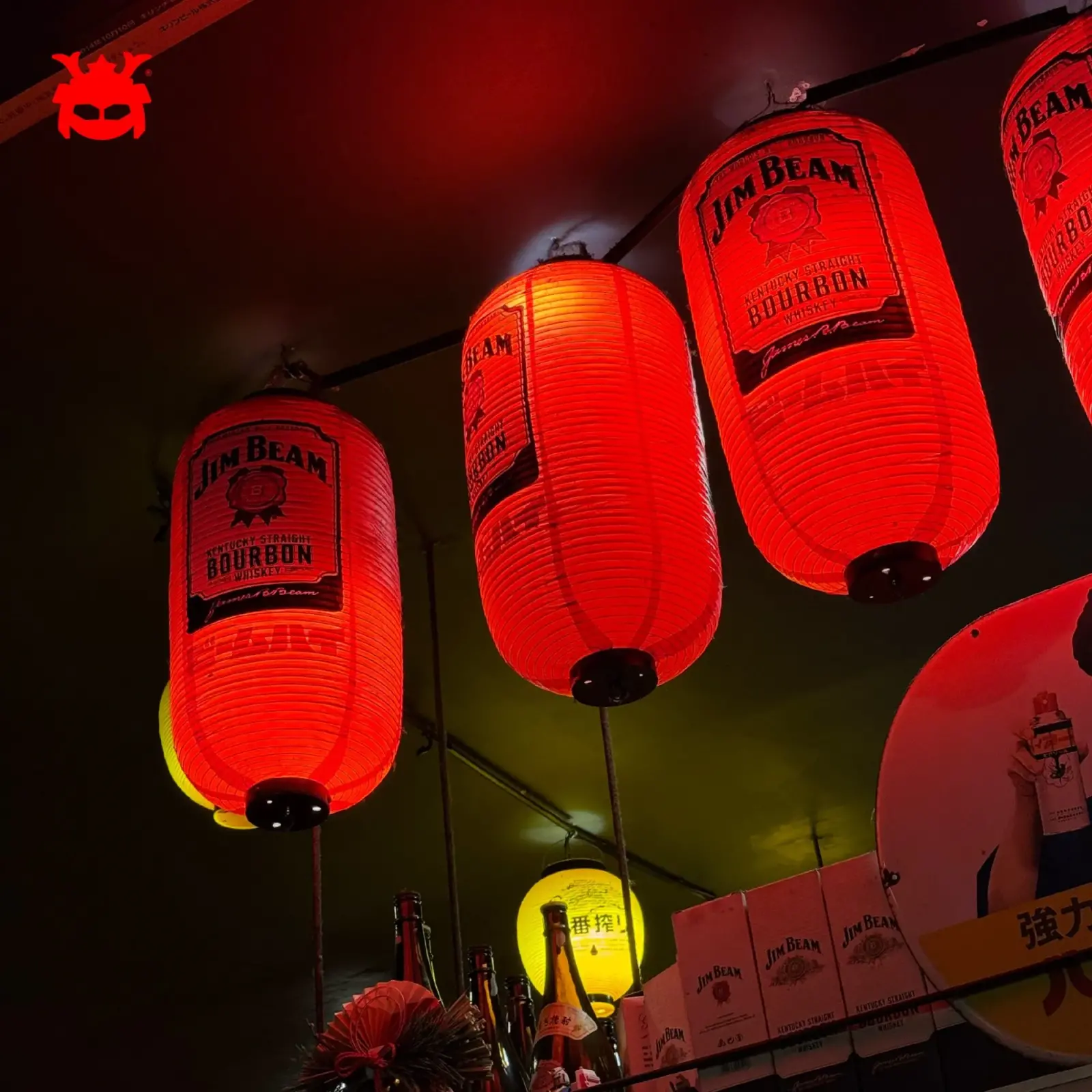 โคมแดงญี่ปุ่นพิมพ์ลาย Jim Beam Bourbon เรียงเป็นแถวเปล่งแสงสีแดงในร้านอิซากายะ